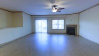 This two-floor property offers a well-designed layout with thoughtful architectural details throughout. The main living area is a spacious, open room highlighted by smooth hardwood-style flooring that reflects natural light generously streaming in from multiple windows. A ceiling fan with a built-in light fixture is centrally mounted, providing both airflow and ambient illumination. Crown molding trims the upper walls, adding a refined finishing touch to the space.

A tiled fireplace with a classic surround serves as a natural focal point along one wall, offering both aesthetic appeal and functional warmth. Adjacent to the fireplace, a standard window frames views of the surrounding neighborhood. On the opposite side of the room, a sliding glass door fitted with vertical blinds opens to what appears to be an outdoor balcony, allowing abundant natural light to fill the interior and providing a seamless connection to the outdoors.

The overall ambiance of the living area is bright, clean, and open, with neutral wall tones and light flooring contributing to an airy, inviting atmosphere. The two-floor layout suggests additional living and private spaces on the upper level, offering a functional separation between common and personal areas.