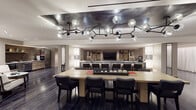 This single-floor space is thoughtfully designed with a contemporary aesthetic, seamlessly integrating several distinct functional zones within an open, flowing layout. Dark hardwood flooring runs throughout, anchoring the refined interiors and providing a consistent visual thread across each area.

The central lounge area is a standout feature, defined by an expansive cognac leather sectional sofa adorned with layered accent pillows in muted tones. A sculptural, branching chandelier with globe glass fixtures serves as the focal point overhead, casting warm, ambient light across the space. Adjacent to the lounge, a long marble-topped bar with dark upholstered seating provides a refined gathering point, complemented by built-in shelving and a large mounted display screen.

The dining area features a substantial light wood table surrounded by black leather chairs beneath the same striking chandelier. A well-appointed kitchenette offers stainless steel appliances, a marble countertop, wood cabinetry, and a textured mosaic tile backsplash.

Quieter corners of the space include a round table with upholstered chairs for intimate meetings, and a minimalist sitting nook featuring sculptural lounge chairs — ideal for private conversation or relaxed retreat.