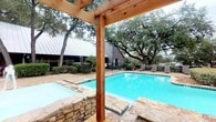 This single-story commercial property combines practical interior office spaces with a well-appointed outdoor amenity area. The exterior features a large, rectangular swimming pool surrounded by a stone and concrete deck, with lounge chairs positioned for relaxation. A rustic wooden pergola with natural timber posts frames the pool area, while mature oak trees provide generous shade and a lush, natural backdrop. A metal-roofed building with gray siding is visible in the background, complementing the landscaped setting enclosed by a black iron fence.

Inside, the office interiors are designed for functionality and professional use. One area features warm wood cabinetry with upper and lower storage units set against light blue walls and carpeted flooring, illuminated by recessed drop-ceiling lighting. An adjacent room, visible through a dark wood-framed doorway, contains built-in shelving and workspace surfaces, suggesting administrative or clerical use.

Additional rooms feature neutral warm-beige walls, drop-tile ceilings with flush lighting panels, and built-in counter surfaces suited for workstation setups. Flooring transitions between carpet and durable vinyl tile depending on the room, supporting a variety of professional or clinical functions. Overall, the space offers a versatile layout that balances indoor professional utility with an attractive and serene outdoor environment.