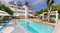 This property at 3720 Tenth Ave in San Diego's Hillcrest area showcases a blend of modern design and resort-style amenities. The complex features multiple stories of residential units with clean, geometric lines and a mix of white and pastel-colored exteriors. Large windows punctuate the façade, suggesting bright, airy interiors. The centerpiece of the property is an expansive pool area, boasting a crystal-clear, uniquely shaped pool surrounded by ample deck space. Lush landscaping, including mature trees and tropical plants, creates a serene oasis-like atmosphere. The pool area is equipped with comfortable lounge chairs, umbrellas for shade, and a pergola-covered seating area, perfect for relaxation or socializing. Additional amenities include a BBQ area and various outdoor seating options. The property's design emphasizes a seamless indoor-outdoor living experience, typical of Southern California's desirable climate and lifestyle.