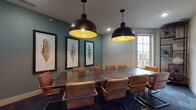 Located at 9815 Seasons West Dr. in Indianapolis, Indiana, this single-floor amenity space blends modern functionality with rustic design touches to create a polished and inviting environment. The centerpiece is a spacious conference room featuring a large industrial-style table surrounded by tan leather chairs, set against teal-painted walls adorned with a trio of framed botanical artwork. Two oversized black dome pendant lights with gold interiors hang above, casting a warm glow, while a generous window allows natural light to fill the room with views of the surrounding community. Patterned carpet tiles in muted blue and gray tones run throughout the space, tying the rooms together cohesively. A distinctive rustic sliding barn door serves as a charming architectural accent, connecting areas with effortless style. The business center offers a dedicated workstation area with multiple computer monitors lined along a wooden desk, illuminated by Edison-style pendant lights against a textured gray feature wall. Warm wood-framed frosted glass doors provide privacy between zones while maintaining an open, connected feel. The overall ambiance is professional yet welcoming, making this space well-suited for meetings, focused work, and collaborative gatherings.