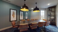 Located at 9815 Seasons West Dr. in Indianapolis, Indiana, this single-floor amenity space blends modern functionality with rustic design touches to create a polished and inviting environment. The centerpiece is a spacious conference room featuring a large industrial-style table surrounded by tan leather chairs, set against teal-painted walls adorned with a trio of framed botanical artwork. Two oversized black dome pendant lights with gold interiors hang above, casting a warm glow, while a generous window allows natural light to fill the room with views of the surrounding community. Patterned carpet tiles in muted blue and gray tones run throughout the space, tying the rooms together cohesively. A distinctive rustic sliding barn door serves as a charming architectural accent, connecting areas with effortless style. The business center offers a dedicated workstation area with multiple computer monitors lined along a wooden desk, illuminated by Edison-style pendant lights against a textured gray feature wall. Warm wood-framed frosted glass doors provide privacy between zones while maintaining an open, connected feel. The overall ambiance is professional yet welcoming, making this space well-suited for meetings, focused work, and collaborative gatherings.