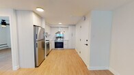 This single-floor unit offers a thoughtfully renovated interior finished with light wood laminate flooring that runs consistently throughout every room, creating a warm and cohesive aesthetic. The kitchen is a standout feature, outfitted with flat-front cabinetry in a crisp white finish, white subway tile backsplash, clean white countertops, and a full suite of stainless steel appliances including a refrigerator, electric range with a stainless steel range hood, and dishwasher. The layout makes efficient use of space while maintaining a clean, modern look. The bathroom continues the contemporary theme with a floating white vanity, matte black fixtures and hardware, white subway tile surrounding the tub-shower combination, and a well-lit, functional layout. The living area and bedroom are open, airy spaces bathed in natural light from large sliding windows that frame views of the surrounding urban neighborhood and cityscape beyond. Baseboard heating runs along the exterior walls beneath each window, ensuring year-round comfort. The overall ambiance of the unit is bright, minimal, and move-in ready, reflecting a clean modern design sensibility throughout.