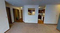 This single-floor space offers a straightforward and functional layout designed for comfortable everyday living. The living area is finished with wall-to-wall carpet, providing a warm and soft underfoot feel, while white walls keep the space feeling bright and clean. A textured ceiling adds a subtle decorative element throughout.

A defining architectural feature is the pass-through window built into the dividing wall between the living area and the kitchen. This opening creates a visual connection between the two spaces without fully opening the floor plan, allowing for easy communication and convenience.

The kitchen is equipped with dark espresso-toned cabinetry that provides a strong contrast against the white walls and appliances. White appliances, including a refrigerator, stove, and dishwasher, are neatly integrated into the cabinetry. The kitchen features hard-surface flooring, offering a practical, easy-to-clean surface. A single-basin sink is positioned along the counter, and under-cabinet lighting provides functional task illumination.

A hallway visible from the living area leads to additional rooms, hinting at a private sleeping and bathing area beyond. The overall ambiance is modest, clean, and functional.