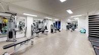 Located at 1717 E Capitol St NE in Washington, DC, this fitness center offers a thoughtfully designed, single-floor workout facility built to accommodate a wide range of exercise routines. The space features a durable speckled epoxy floor throughout, providing a clean and resilient surface ideal for high-traffic gym use. Bright recessed fluorescent lighting illuminates the entire area evenly, creating a well-lit environment conducive to focused training.

The strength training section is outfitted with a variety of cable and resistance machines arranged in an open, easy-to-navigate layout. Full-length mirrors line the walls, enhancing the sense of spaciousness while allowing users to monitor their form. The cardio zone includes a row of treadmills and elliptical machines, each equipped with individual monitor screens for entertainment during workouts.

A dedicated functional fitness area features medicine balls in assorted sizes, stability balls, resistance tools, and free weight racks, supporting dynamic and versatile training sessions. A striking horizontal slat accent wall in dark wood tones adds a bold design element, offering a visual contrast to the otherwise clean white walls and structural columns. The overall ambiance is professional, organized, and inviting, making this fitness center a comprehensive resource for residents and users in the Capitol Hill neighborhood.