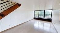 This two-floor space presents a clean, open layout with a minimalist aesthetic. The main level is characterized by polished terrazzo flooring that reflects natural light, contributing to a bright and airy atmosphere. White walls provide a neutral, versatile backdrop throughout, offering a blank canvas suitable for a variety of uses.

A large, multi-pane window with a dark metal frame anchors one wall, inviting abundant natural light and framing views of surrounding greenery. A baseboard heating unit runs beneath the window, providing efficient climate control. The exposed wooden staircase, featuring a substantial solid wood beam, introduces a warm, rustic contrast to the otherwise contemporary, pared-back interior, and serves as a notable architectural element within the space.

The white walls show subtle paneling details, adding a degree of texture without disrupting the clean visual aesthetic. The overall ambiance is functional and uncluttered, making this two-floor space well-suited for creative, professional, or flexible-use purposes. The combination of durable flooring, generous natural light, and a straightforward layout provides a practical and adaptable environment.