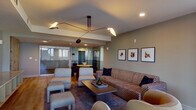 This thoughtfully designed single-floor space offers a seamless blend of contemporary style and functional layout. The main living area serves as the centerpiece, anchored by a striking multi-arm sculptural pendant light suspended from a coffered ceiling. A generously sized patterned sofa, coordinating accent chairs, upholstered ottomans, and a ribbed wood coffee table create a comfortable, curated seating arrangement over a layered area rug atop warm hardwood flooring that extends throughout.

A glass-walled room adjacent to the living area adds architectural interest and visual depth, offering transparency and a sense of openness. Framed artwork lines the walls, contributing a gallery-like quality to the space. A round dining table with dark chairs occupies a bright corner nearby.

A separate dining area features expansive curved floor-to-ceiling windows on multiple sides, flooding the room with natural light and offering sweeping city skyline views. This area also provides direct access to an outdoor terrace, further enhancing the connection to the surrounding urban landscape.

A galley-style kitchen corridor showcases distinctive textured tile accent walls, light wood cabinetry, and a continuous countertop surface, adding warmth and a refined material contrast. Throughout, the space maintains a cohesive, modern aesthetic with carefully considered finishes and furnishings.