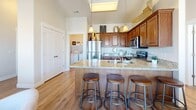 Located at 1037 W River Fox Way in Midvale, Utah, this well-appointed one-bedroom, one-bathroom residence offers a thoughtfully designed single-floor layout. Warm hardwood-style flooring flows throughout the main living areas, anchoring an open-concept living and dining space filled with natural light from large windows dressed with elegant drape panels. A ceiling fan adds comfort to the inviting living room, which offers views of the surrounding neighborhood. The dining area is tucked alongside a glass door, enhancing the sense of openness and providing easy outdoor access. The kitchen is a standout feature, boasting rich wood cabinetry, granite countertops, a spacious island with bar seating for three, stainless steel appliances, and soaring ceilings with pendant lighting that amplify the airy feel of the space. A dedicated laundry area with washer and dryer is conveniently accessible from the kitchen. The generously sized bedroom offers plush carpeting, a ceiling fan, and access to a walk-in closet with built-in shelving and hanging rods, complemented by two additional closets for ample storage. The bathroom features a full tub-shower combo. Three total closets ensure outstanding storage throughout this efficiently designed home.