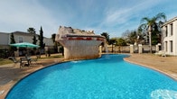 This inviting residential community is designed around an open-air courtyard layout, offering a cohesive and social living environment. The grounds are anchored by a striking freeform swimming pool with crystal-clear blue water, whose most distinctive feature is a large, sculptural stone waterfall centerpiece — an eye-catching architectural element that cascades water into the pool below. The pool area is surrounded by a paved deck and complemented by shaded seating with patio furniture and umbrellas, creating an ideal outdoor retreat.

The buildings themselves are arranged around a central courtyard, with units accessed via covered exterior walkways and staircases fitted with black metal railings. Each unit entry features a bold red door set against white-painted siding, offering a classic and welcoming aesthetic. The tiled walkways running along the building facades provide direct access to individual units while maintaining an open, airy feel.

Throughout the property, palm trees and manicured greenery add a subtropical character to the landscaping, enhancing the warm, resort-like atmosphere. The courtyard layout allows for natural light to flow throughout the common areas, and the elevated walkways offer pleasant views over the grounds. The overall ambiance is casual, sunny, and community-oriented, making this a comfortable and characterful place to call home.