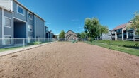 This outdoor dog park is a dedicated pet amenity situated within a multi-building residential community. The park features a generously sized, open sandy ground surface that provides ample room for pets to run and play freely. A secure chain-link fence encloses the entire perimeter, with a gated entry point for safe and easy access. Inside the park, a green agility ramp offers an engaging obstacle for active dogs of all sizes.

The space is framed by mature deciduous trees that provide natural shade and a pleasant, park-like atmosphere. Surrounding the dog park are well-maintained green lawns and paved walkways that connect the various residential buildings, which feature gray and blue horizontal siding and multi-story layouts. The open-air setting benefits from abundant natural light under clear skies, creating an inviting environment for both pets and their owners.

The grounds beyond the fencing include additional landscaped lawn areas, street lighting, and parking, offering a well-rounded community outdoor experience. The overall setting is clean, functional, and thoughtfully designed to serve residents seeking a safe and accessible space for their pets to exercise and socialize.