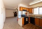 Located at 10301 Golf Course Rd NW in Albuquerque, New Mexico, this single-story residence offers a practical and comfortable layout suited for everyday living. The kitchen is well-appointed with warm wood-toned cabinetry, a black gas range with overhead ventilation, a black dishwasher, and a double-basin sink beneath a window that brings in natural light and views of the surrounding landscape. A peninsula counter provides a convenient pass-through to the adjoining living area, enhancing the open feel of the space. Wood-look flooring runs through the kitchen, while plush carpeting extends throughout the living room and bedrooms. The spacious living room is highlighted by a corner fireplace and a ceiling fan, with sliding glass patio doors that open to an outdoor view, filling the room with natural light. The bedrooms are generously sized, each featuring mirrored sliding closet doors and windows that overlook the exterior greenery. Two full bathrooms include tub-shower combinations with tiled surrounds, wood-toned vanities with granite-style countertops, and wood-look flooring. A dedicated hallway connects the bedrooms and bathrooms, adding to the functional flow of the home.