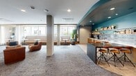 Located at 201 The Promenade N in Long Beach, California, this single-floor space offers a generously sized, thoughtfully designed communal environment. The layout flows from wide, light-filled hallways finished with large-format wood-look tile flooring into an expansive open-plan kitchen and living area. The transition between zones is marked by a shift to patterned carpet, adding texture and visual warmth underfoot.

The living area is anchored by plush cognac leather lounge chairs and built-in banquette seating upholstered in neutral fabric, arranged along floor-to-ceiling windows that face the surrounding streetscape, flooding the space with natural light. Sleek pedestal side tables accompany the seating throughout. Pendant lighting with an industrial character hangs at varying heights, complementing recessed ceiling fixtures overhead.

A kitchen counter with a dark stone surface and bar-height stools defines the culinary zone, backed by teal-painted walls, open wood shelving, and decorative tile. The bold teal ceiling accent with integrated LED strip lighting creates a dramatic visual boundary between the kitchen and lounge areas.

Black steel-framed sliding barn doors provide access to a private room or flex space, adding an architectural focal point. Curated artwork and indoor plants contribute to an inviting, design-forward ambiance throughout.