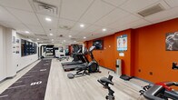 Located at 16 W King St in Lancaster, PA, this health club offers a comprehensive fitness facility within a single, efficiently organized floor. The space is designed with a long, rectangular layout that maximizes its footprint, accommodating a diverse range of workout stations without feeling congested.

The interior features light wood-style flooring throughout, contrasted by bold orange accent walls that lend the space an energetic, motivating atmosphere. A drop-panel ceiling with recessed lighting provides consistent, bright illumination across the entire floor. A central turf track runs the length of the room, marked with yardage lines, suitable for sprint drills and functional training.

Cardio equipment lines one side of the space and includes treadmills, elliptical trainers, a stationary bike, and a rowing machine. On the opposite end, a dedicated strength and functional training zone features a cable machine, adjustable bench, medicine balls, kettlebells, resistance bands, and a punching bag. Large mirrors along one wall enhance the sense of depth. A wall-mounted flat-screen TV is positioned for visibility across the floor. Windows along one wall allow natural light to supplement the overhead lighting, contributing to an open, welcoming environment.