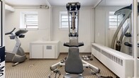 Located at 1040 Hollington St in Ottawa, Ontario, this exercise room offers a well-equipped fitness space designed to support a range of workout routines. Despite its compact footprint, the room is thoughtfully arranged to maximize functionality and usability.

The space is furnished with a multi-function cable strength machine, two upright stationary bicycles — including a Life Fitness model — and a set of free weights, providing options for both strength training and cardiovascular exercise. A large floor-to-ceiling mirror along one wall visually expands the room and supports proper form during workouts.

Natural light filters in through multiple windows equipped with blinds, lending the space a bright and welcoming atmosphere. The neutral-toned carpet adds a layer of comfort underfoot, while a baseboard heater ensures the room remains usable year-round in Ottawa's variable climate. Built-in white cabinetry provides convenient storage.

A striking black-and-white graphic mural dominates one wall, incorporating local references including the address "1040 Hollington" and a Canadian flag, adding a vibrant, identity-rich character to the room. Exposed piping along the ceiling adds an industrial aesthetic that complements the mural's urban style. The space is accessed through a standard door and is clearly designated as a no-smoking area.