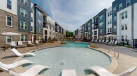 Located at 9111 Paragon Dr in Charlotte, NC, this outdoor amenity space is designed to deliver a resort-like experience within a contemporary residential community. At its centerpiece is a generously sized freeform swimming pool with crystal-clear water, gradual entry points, and stainless steel handrails. The pool is surrounded by a wide, smooth concrete sundeck lined with lounge chairs, umbrellas, and Adirondack-style seating, providing ample space for relaxation and recreation. Thoughtfully designed landscaping features ornamental grasses, mulched planting beds, and manicured greenery, adding natural texture and color throughout the space. Black iron perimeter fencing defines the pool area while maintaining an open, airy feel. A covered pergola structure is visible adjacent to the pool deck, offering a shaded gathering area. The backdrop consists of a modern multi-story residential building clad in a combination of brick, navy blue, white, and gray siding with private balconies. A separate clubhouse-style building is also visible, complementing the overall architectural aesthetic. The entire outdoor area is thoughtfully illuminated with bollard-style lighting, ensuring functionality and ambiance during evening hours. The space is open to the sky, inviting abundant natural light throughout the day.