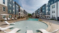 Located at 9111 Paragon Dr in Charlotte, NC, this outdoor amenity space is designed to deliver a resort-like experience within a contemporary residential community. At its centerpiece is a generously sized freeform swimming pool with crystal-clear water, gradual entry points, and stainless steel handrails. The pool is surrounded by a wide, smooth concrete sundeck lined with lounge chairs, umbrellas, and Adirondack-style seating, providing ample space for relaxation and recreation. Thoughtfully designed landscaping features ornamental grasses, mulched planting beds, and manicured greenery, adding natural texture and color throughout the space. Black iron perimeter fencing defines the pool area while maintaining an open, airy feel. A covered pergola structure is visible adjacent to the pool deck, offering a shaded gathering area. The backdrop consists of a modern multi-story residential building clad in a combination of brick, navy blue, white, and gray siding with private balconies. A separate clubhouse-style building is also visible, complementing the overall architectural aesthetic. The entire outdoor area is thoughtfully illuminated with bollard-style lighting, ensuring functionality and ambiance during evening hours. The space is open to the sky, inviting abundant natural light throughout the day.
