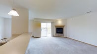 This single-floor space centers around a spacious, open living area finished with plush wall-to-wall carpeting in a neutral tone that complements the light-colored walls and creates a bright, airy atmosphere. A standout feature of the room is the corner fireplace, framed by a stone or tile surround and topped with a warm wood mantel, adding both a functional heating element and a focal point to the space. Adjacent to the fireplace, a sliding glass door fills the room with natural light while providing direct access to an outdoor area and views of the surrounding exterior. A half-wall or countertop partition is visible along one edge of the space, suggesting a transition to a kitchen or dining area and offering a practical divide without fully closing off the open layout. Recessed or spot lighting supplements the natural light, ensuring the interior remains well-lit. The overall design is clean and understated, with a neutral palette throughout that lends itself well to a variety of furnishing styles. The layout feels open and functional, making efficient use of the available floor space on this single level.