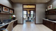 This refined single-floor professional space is thoughtfully designed to support both collaboration and productivity. The interior is unified by a consistent design language featuring textured grid-patterned wall coverings, rich dark wood trim and cabinetry, light wood-plank flooring, and granite countertops that lend an air of understated elegance throughout.

The centerpiece of the space is a well-appointed conference room, accessed through a framed opening with a transom window above. The room accommodates a large dark wood conference table surrounded by ergonomic white leather chairs. Natural light filters in through tall windows dressed with layered roman shades and floor-length drapes, offering views of lush outdoor greenery. A pendant chandelier with a drum shade anchors the room's ceiling.

Adjacent to the conference room is a comfortable lounge area featuring white upholstered armchairs, dark wood shelving with decorative accents, and a striking large-scale abstract artwork in warm amber and red tones that serves as a bold focal point on the wall.

A dedicated business center workstation includes a built-in granite-topped desk with dark wood cabinetry below, a desktop computer, printer, and a framed display shelf above — providing a functional workspace within the suite.

The main entry area features glass French doors with transom windows, flooding the space with natural light and providing a welcoming, open feel upon arrival.