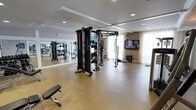 This fitness facility is thoughtfully designed to accommodate a comprehensive workout experience within a single-floor layout. The space features durable, light-toned flooring that provides both functionality and a clean aesthetic throughout the room. A large mirrored wall runs along one side, visually expanding the room and allowing users to monitor their form during exercise.

At the center of the space stands a multi-functional cable and resistance training rig, offering a wide range of strength and conditioning exercises. An adjustable workout bench is positioned nearby, complementing the available equipment. Additional free weights and accessories are visible along the mirrored wall, providing further versatility for training routines.

Recessed ceiling lighting illuminates the space evenly, while sheer curtains along the windows allow natural light to filter in, contributing to a bright and open atmosphere. A wall-mounted flat-screen television is installed on one end of the room, enabling users to follow workout programs or entertainment during sessions. The overall design balances practicality with a clean, modern aesthetic, creating an inviting environment for fitness activities.