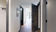 Located at 401 E State St in downtown Ithaca, this 2-bedroom, 1-bathroom residence showcases a striking industrial-modern aesthetic across a single floor of well-utilized living space. The unit's architectural character is defined by soaring ceilings with exposed ductwork and structural elements, paired with large, multi-pane factory-style windows that flood the interior with natural light and frame views of the surrounding cityscape.

The open-concept kitchen and living area serves as the centerpiece of the unit. The chef's kitchen features a standout curved, fluted-panel island with a dramatic veined stone countertop, complemented by sleek slate-blue cabinetry, stone counters along the perimeter, white tile backsplash, stainless steel appliances, and a globe pendant light fixture. The generous living area benefits from expansive windows along an entire wall, providing abundant daylight and outdoor views.

Both bedrooms feature light wood-tone flooring and clean white walls, with each room accented by bold, dark-framed doors and trim. The bathroom is finished with large-format gray tile flooring, a wide vanity with a white countertop, and matte black fixtures. The unit also includes two closets and in-unit laundry, offering practical, well-organized living in a distinctive, design-forward space.