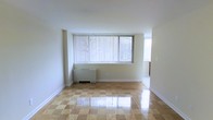 This single-floor space offers a clean, functional layout with a bright and airy atmosphere. The main living area is immediately welcoming, characterized by its eye-catching polished checkered tile flooring that adds a classic, timeless charm to the interior. Large windows dressed with vertical blinds dominate one wall, flooding the space with generous natural light and offering views of the surrounding greenery outside. The neutral white walls and white baseboards create a fresh, versatile backdrop that complements a variety of décor styles. A wall-mounted radiator unit is positioned beneath the main window, providing efficient in-unit heating. The open layout of the living area provides a sense of spaciousness and flexibility for furniture arrangement. A secondary window is visible along the adjacent wall, further enhancing the natural brightness of the room. The overall ambiance of the space is light, clean, and uncluttered, making it a welcoming and adaptable environment suited for comfortable everyday living.