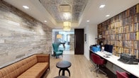 This single-floor space makes a strong design statement through its thoughtful blend of rustic and contemporary elements. A striking reclaimed wood accent wall anchors the lounge area, where a striped sofa and a teal wingback chair create an inviting seating arrangement centered around a round pedestal side table. An ornate tin-tile ceiling with an embossed metallic finish adds architectural character overhead, complemented by a decorative pendant light fixture that serves as a focal point.

The space transitions naturally into a dedicated work area, where a built-in shelving unit filled with books lines the wall alongside a functional desk setup with seating. The workspace is equipped with a computer and office peripherals, making it both practical and stylish. Light wood flooring runs throughout, unifying the two zones cohesively.

A glass-partitioned area in the background offers a glimpse into an adjacent room, enhancing the sense of depth and openness. Recessed lighting supplements the natural and decorative light sources, ensuring the space is well-lit throughout. The overall ambiance is warm, eclectic, and professionally curated.