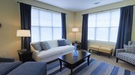 This thoughtfully designed single-floor space offers a comfortable and inviting atmosphere highlighted by an elegantly appointed living area. The living room is a standout feature, bathed in abundant natural light streaming through multiple large windows dressed with crisp white blinds and rich navy blue curtains that add a sophisticated touch to the room. The soft, neutral-toned walls complement the carefully chosen furnishings, including a plush white sofa adorned with textured throw pillows in shades of blue and gray. A dark wood coffee table anchors the seating area, accented by a striking blue decorative vase that adds a pop of color. A woven blue-toned area rug defines the space and ties the color scheme together seamlessly. Additional seating is provided by a classic upholstered bench, while a floor lamp and side table offer both functional lighting and decorative charm. The room's layout promotes an open, airy feel, making it well-suited for relaxing or entertaining. Carpeted flooring throughout the living area adds warmth and comfort underfoot. Overall, the space presents a clean, modern aesthetic with a welcoming ambiance defined by its harmonious blend of natural light, neutral tones, and coordinated navy accents.
