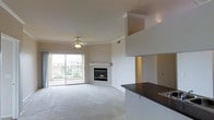 This single-floor space features an open-concept layout that seamlessly connects the living area and kitchen. The living room is finished with plush wall-to-wall carpeting and is anchored by a gas fireplace set into a clean white surround, adding warmth and a focal point to the room. Crown molding lines the upper walls, lending an elegant architectural detail throughout. A ceiling fan with a light fixture is centrally positioned, providing both air circulation and ambient lighting. Large sliding glass doors flood the space with natural light and lead to a private balcony, offering views of the surrounding neighborhood. The transition to the kitchen is defined by a raised countertop bar with a dark granite or stone surface, creating a natural division between the two areas while maintaining the open feel. The kitchen features a sink integrated into the counter. The walls throughout are finished in neutral tones, complementing the light-colored carpet and white trim. The overall ambiance is clean, airy, and well-appointed, with thoughtful details that contribute to a comfortable and functional living environment.