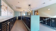 Located at 2603 North Fry Road in Katy, Texas, this mail room serves as a functional and thoughtfully designed communal hallway space. The room is lined on all sides with rows of numbered black metal mailboxes, providing organized mail retrieval for a large number of residents. A central island unit extends along the length of the hallway, maximizing mailbox capacity while maintaining a clear pathway for easy navigation.

The interior design is cohesive and polished, featuring soft sage-green walls that lend a calm, welcoming atmosphere. Overhead, distinctive cluster pendant lights with exposed Edison-style bulbs hang from the ceiling, adding a contemporary, decorative touch to an otherwise utilitarian space. The neutral-toned flooring complements the overall color palette and contributes to the room's clean aesthetic.

Framed abstract artwork adorns one wall, elevating the space beyond a standard utility area and giving it a more refined character. Natural light filters in through a window visible at one end of the hallway, brightening the interior. A wall-mounted sign clearly identifies this as the mail room for 2603 N. Fry Road, and a sleek waste receptacle is conveniently positioned near the entrance. The space is efficiently organized, balancing practicality with an appealing, modern design sensibility.