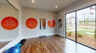 This single-floor fitness and yoga studio is thoughtfully designed to support a range of wellness activities. The space features warm hardwood flooring throughout, providing a durable and attractive surface well-suited for yoga, stretching, and group exercise. Recessed lighting in the ceiling ensures consistent illumination across the room, complemented by abundant natural light streaming in through large sliding glass doors that offer views of an outdoor landscaped area.

A full wall of mirrors amplifies the sense of space and allows users to monitor their form during workouts. The walls are adorned with bold, orange circular graphics featuring motivational quotes, including one attributed to Rumi, adding an inspirational and energetic ambiance. A teal "Namaste" script decal reinforces the studio's wellness-focused identity.

The studio is well-stocked with equipment, including stability balls in multiple sizes, wall-mounted yoga mat racks holding rolled orange mats, a black exercise mat, medicine balls on a rack, resistance bands, step platforms, and suspension training accessories. The clean, neutral wall tones provide a calm backdrop that balances the vibrant décor, creating an environment that is both motivating and conducive to focused practice.