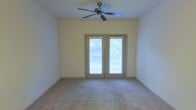 This single-story space features a well-proportioned main room finished with wall-to-wall carpeting in a neutral tone, contributing to a clean and understated aesthetic. The walls are painted in a soft, light cream color, creating a bright and airy atmosphere throughout. A ceiling fan with classic dark-finished blades is centrally mounted, providing both functional air circulation and a modest architectural focal point overhead. The room's most notable feature is a set of French doors framed in a complementary neutral trim, fitted with horizontal blinds that allow for adjustable light control. These doors open to an outdoor balcony, offering views of surrounding greenery, which enhances the sense of connection to the outdoors and brings natural light into the interior. The balcony railing is visible through the glass panels, suggesting a comfortable outdoor extension of the living space. The overall design is straightforward and functional, with a neutral color palette throughout that offers versatility for a variety of furnishing styles. The combination of soft carpeting, natural light, and outdoor access makes this a comfortable and practical living area.