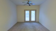 This single-story space features a well-proportioned main room finished with wall-to-wall carpeting in a neutral tone, contributing to a clean and understated aesthetic. The walls are painted in a soft, light cream color, creating a bright and airy atmosphere throughout. A ceiling fan with classic dark-finished blades is centrally mounted, providing both functional air circulation and a modest architectural focal point overhead. The room's most notable feature is a set of French doors framed in a complementary neutral trim, fitted with horizontal blinds that allow for adjustable light control. These doors open to an outdoor balcony, offering views of surrounding greenery, which enhances the sense of connection to the outdoors and brings natural light into the interior. The balcony railing is visible through the glass panels, suggesting a comfortable outdoor extension of the living space. The overall design is straightforward and functional, with a neutral color palette throughout that offers versatility for a variety of furnishing styles. The combination of soft carpeting, natural light, and outdoor access makes this a comfortable and practical living area.