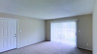 This single-floor space presents a straightforward and functional layout highlighted by a well-proportioned living area. The room features wall-to-wall carpeting in a neutral beige tone, contributing to a warm and understated aesthetic. A prominent sliding glass door serves as the focal point of the main living space, fitted with full-length vertical blinds that allow for flexible light control. When open, the blinds reveal a view of outdoor greenery, bringing a natural element into the interior. The walls are finished in a soft, neutral gray-beige tone that complements the flooring and creates a cohesive, understated palette throughout. The ceiling features a classic textured popcorn finish and is set at a standard height, maintaining a comfortable sense of enclosure. White baseboards provide clean contrast against the walls and carpet. The overall ambiance is calm and move-in ready, offering a blank canvas well-suited for a variety of furniture arrangements and personal styling preferences. The space is practical in its design, prioritizing openness and ease of use.