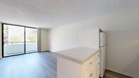 Located at 606 Third Avenue in San Diego, CA, this efficiently designed single-floor unit offers a well-organized layout comprising a living room, kitchen, hallway, and full bathroom. The open-plan living and kitchen area is anchored by a peninsula with granite-style countertops and white cabinetry, providing both storage and a natural division between cooking and living spaces. The kitchen is equipped with a stove, microwave, and refrigerator, all finished in a clean, white palette that complements the bright interior. Light wood-toned laminate flooring runs throughout the unit, creating a cohesive and warm aesthetic. The living area is bathed in natural light through a large sliding glass door that opens onto a private balcony, offering views of the surrounding urban greenery. A textured ceiling adds a classic residential touch overhead. The hallway connects the main living space to the bathroom and entry, and features built-in cabinetry for additional storage. The bathroom includes a full shower-tub combo with white tile surround and chrome fixtures. The unit's neutral color palette of soft beige and white tones throughout creates a clean, inviting atmosphere well-suited to the vibrant downtown San Diego location.