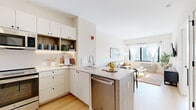 Located at 215 Grove St in Jersey City, NJ, this thoughtfully designed one-bedroom, one-bathroom residence offers a modern aesthetic with clean lines and a bright, neutral palette throughout. Light hardwood flooring flows seamlessly across the entire floor plan, enhancing the sense of openness and continuity.

The open-concept kitchen is a standout feature, outfitted with white flat-front cabinetry, warm wood accent shelving with under-cabinet lighting, quartz countertops, and a full suite of stainless steel appliances including a refrigerator, range, microwave, and dishwasher. A peninsula with an integrated sink provides additional prep space and connects effortlessly to the living area.

The living room is bathed in natural light through large windows that frame impressive city skyline views, creating an inviting and airy atmosphere. The bedroom offers a peaceful retreat with its own city-facing window, ample natural light, and a cozy, well-proportioned layout. 

The bathroom is finished with full-height white subway tile surrounding a soaking tub and shower combination, complemented by a floating vanity with a large illuminated mirror and neutral tile flooring, delivering a clean, spa-inspired feel. The overall design of this residence balances modern functionality with a warm, understated elegance.