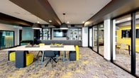 Located at 1910 S Union St in Anaheim, California, this single-floor commercial space delivers a striking, contemporary design built for collaboration and productivity. The interior is defined by a bold color palette of vivid yellow, deep charcoal, and cool gray, carried consistently throughout the space via statement furniture, patterned carpeting, and accent walls.

The main gathering area features a large, light-toned communal table surrounded by a mix of yellow and dark upholstered chairs, ideal for team meetings or focused work sessions. Dramatic dome pendant lights hang overhead, complementing the dark ceiling treatment that adds depth and sophistication. A built-in shelving unit with a wall-mounted television serves as a functional focal point along one wall, flanked by a beverage station and decorative display shelving.

A separate lounge zone offers casual seating with low-profile yellow accent chairs and a compact coffee table, bathed in natural light from floor-to-ceiling glass walls that overlook the exterior. A teal-painted ceiling detail adds a pop of color to this relaxed nook.

A glass-partitioned private conference room provides an intimate setting for smaller meetings, featuring a wall-mounted display, a sculptural geometric pendant light, and a vivid yellow accent wall. Throughout, the patterned carpet ties the design language together, creating a cohesive, energetic, and professional atmosphere.