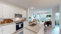 This single-floor residence at The Statesman showcases a thoughtfully designed open-concept layout with contemporary finishes throughout. The kitchen serves as a central hub of the living space, featuring white shaker cabinetry extending to the ceiling, granite countertops, a subway tile backsplash, and a full suite of stainless steel appliances including a French door refrigerator, range, and built-in microwave. A large central island with a single-basin sink and bar seating provides additional prep and dining space, while light wood-tone flooring runs seamlessly throughout the main living areas.

The living room is bright and welcoming, benefiting from generous natural light through sliding glass doors that open to a private balcony with outdoor views. A modern chandelier adds an elegant focal point overhead. The bedroom offers a comfortable retreat with multiple windows fitted with plantation shutters, allowing for adjustable natural light. An additional bedroom provides equally well-lit accommodations with outdoor-facing windows.

The bathroom presents a clean, elongated layout with a double-sink granite vanity, wide mirror, and crisp white cabinetry. Throughout the residence, the consistent use of light tones, recessed lighting, and quality materials creates a cohesive, modern ambiance.