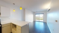 Located at 1629–1655 John Deshields Blvd in Bentonville, Arkansas, this single-floor unit presents a clean, modern design throughout. The open-concept living and dining area is anchored by dark hardwood-style flooring and bright white walls, creating a crisp, contemporary ambiance. A large sliding glass door floods the living space with natural light and provides direct views of the surrounding greenery and streetscape beyond.

The kitchen is well-appointed with stainless steel appliances, including a side-by-side refrigerator with ice and water dispenser, a stainless range with overhead ventilation, and a dishwasher. White upper cabinetry contrasts with muted sage-toned lower cabinets, complemented by clean white countertops and under-cabinet lighting. Distinctive yellow pendant lights add a warm, accent detail above the kitchen island and dining area.

The bedroom features the same rich hardwood-style flooring, a ceiling fan, a closet, and a window offering natural light and outdoor views. The bathroom is finished with tile flooring, an illuminated vanity mirror, neutral cabinetry, white countertops, and a tiled shower. A stacked washer and dryer unit is conveniently located adjacent to the bathroom, adding practical functionality to the space.