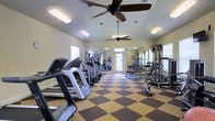 This fitness center offers a spacious and well-organized workout environment designed to support a full range of exercise routines. The room features a distinctive two-tone checkered floor in brown and tan, adding a dynamic visual element to the space. A row of cardio machines — including treadmills and elliptical trainers — lines one side of the room, providing ample options for aerobic exercise. On the opposite side, a dedicated strength training area is equipped with weight machines, free weights, and benches, catering to resistance and muscle-building workouts. Full-length mirrored walls run along the back of the room, visually expanding the space and allowing users to monitor their form. Two ceiling fans are mounted overhead, promoting air circulation throughout the facility and contributing to a comfortable workout environment. Recessed and flush-mounted lighting ensures the room is well-lit at all times. Natural light filters in through multiple windows and a set of glass doors at the far end, offering views of the surrounding outdoor area. The overall ambiance is clean, functional, and inviting, making it a practical and motivating space for fitness enthusiasts.