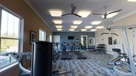 This fitness center is thoughtfully designed to support a full range of workout routines. The space is finished with a geometric, multi-tone patterned flooring that adds visual interest underfoot, while the walls are painted in a neutral tan and a deep blue-gray accent shade, giving the room a clean, contemporary feel. Crown molding along the upper walls adds a refined architectural touch.

The ceiling is outfitted with an array of large, square flush-mount light panels that ensure bright, even illumination throughout the space. Multiple ceiling fans are evenly distributed across the room, promoting consistent air circulation during workouts. Windows along one wall allow natural light to filter in, enhancing the overall brightness of the environment.

The cardio area is well-stocked with a row of treadmills and elliptical machines arranged in a line, accompanied by a wall-mounted flat-screen television for entertainment. A dedicated free weight section features a dumbbell rack along the far wall, paired with a weight bench. A Paramount multi-station strength machine anchors one side of the floor, rounding out the available equipment. A mirrored wall near the back expands the sense of space and supports form-checking during exercise.