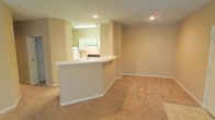 This single-floor space is designed with an open, functional layout that seamlessly connects the kitchen and living areas. The kitchen is partially enclosed, featuring white cabinetry, a stove, and a refrigerator, while a white breakfast bar with a raised countertop serves as a dividing element between the kitchen and the main living space. Recessed lighting and an under-cabinet fluorescent fixture illuminate the kitchen area, keeping it bright and practical. The living area is finished with wall-to-wall beige carpeting, providing a warm and comfortable surface underfoot. Neutral beige walls run throughout the space, creating a consistent and understated aesthetic that lends itself well to a variety of interior styles. White baseboards trim the perimeter of the room, adding a clean, finished look. The open floor plan allows for flexible furniture arrangements within the living area, maximizing the usable space. Overall, the design prioritizes simplicity and practicality, making it a versatile and move-in-ready environment.