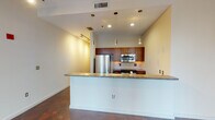 This single-floor urban residence showcases a thoughtfully designed open-concept layout that maximizes both space and functionality. The living area is anchored by striking polished concrete floors that extend throughout, lending an industrial-chic aesthetic to the space. Large floor-to-ceiling windows flood the living area with abundant natural light while framing impressive city skyline views, creating a strong sense of connection to the urban environment outside.

The kitchen is positioned centrally behind a granite-topped breakfast bar that doubles as a divider between the cooking and living spaces. Rich medium-toned wood cabinetry provides ample storage, while the kitchen is equipped with stainless steel appliances including a refrigerator, stove, and built-in microwave. Granite countertops with a stainless steel sink complete the functional and polished workspace. Pendant lighting and recessed ceiling lights illuminate the kitchen area warmly.

Track lighting installed along the ceiling offers flexible illumination throughout the main living space. The bathroom features a full soaking tub with a tiled surround, accented with a decorative mosaic tile band, complemented by tile flooring and a granite-topped vanity. Clean white walls throughout the unit create a bright, neutral backdrop that enhances the overall modern and contemporary character of the space.