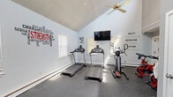 Located at 240 New State Rd in Manchester, CT, this dedicated exercise room offers a well-equipped fitness environment designed to support a variety of cardio workouts. The space features soaring vaulted ceilings that create an open, airy atmosphere, complemented by a ceiling fan that aids in air circulation during workouts. Natural light filters in through multiple windows fitted with white blinds, brightening the room throughout the day. Upper clerestory windows add to the sense of height and openness.

The rubber-matted flooring provides a durable, slip-resistant surface ideal for exercise equipment and free weights. The room is outfitted with two treadmills, an elliptical machine, and spin bikes, catering to a range of cardio preferences. A rack of kettlebells and dumbbells offers options for strength training as well. A wall-mounted flat-screen television ensures entertainment during workouts, and a full-length mirror along one wall expands the sense of space.

Motivational wall decals are a defining design feature, with bold fitness-themed typography — including phrases like "Strength," "Endurance," "Ability," "Motivation," and "Attitude" — adorning the light blue and gray walls. The overall ambiance is energetic and encouraging, making this fitness center a functional and inspiring place to exercise.