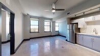 This single-floor unit presents a contemporary urban design characterized by polished concrete floors that reflect natural light throughout the open living space. The main living area is generously proportioned, featuring a ceiling fan, neutral-toned walls with bold dark baseboards, and multiple black-framed windows that frame sweeping elevated city views, flooding the interior with abundant natural light.

The kitchen is seamlessly integrated into the open-plan layout and is well-appointed with white flat-front cabinetry, a white tile backsplash, light countertops, and a full suite of stainless steel appliances including a refrigerator, dishwasher, stainless range, and built-in microwave. Track lighting overhead ensures the workspace is well-lit, and a louvered utility door adds a functional design detail nearby.

A separate bedroom features its own black-framed windows with elevated city views, continuing the consistent design language found throughout the unit. The overall aesthetic is clean and modern, combining industrial elements like polished concrete with refined finishes to create a cohesive, contemporary urban living environment.