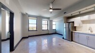 This single-floor unit presents a contemporary urban design characterized by polished concrete floors that reflect natural light throughout the open living space. The main living area is generously proportioned, featuring a ceiling fan, neutral-toned walls with bold dark baseboards, and multiple black-framed windows that frame sweeping elevated city views, flooding the interior with abundant natural light.

The kitchen is seamlessly integrated into the open-plan layout and is well-appointed with white flat-front cabinetry, a white tile backsplash, light countertops, and a full suite of stainless steel appliances including a refrigerator, dishwasher, stainless range, and built-in microwave. Track lighting overhead ensures the workspace is well-lit, and a louvered utility door adds a functional design detail nearby.

A separate bedroom features its own black-framed windows with elevated city views, continuing the consistent design language found throughout the unit. The overall aesthetic is clean and modern, combining industrial elements like polished concrete with refined finishes to create a cohesive, contemporary urban living environment.