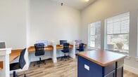 Located at 502 S Main St in Warrensburg, Missouri, this single-room office space offers a well-designed, functional workspace. Despite its compact footprint, the room feels open and airy thanks to notably high ceilings and large windows fitted with horizontal blinds that allow ample natural light to fill the space.

The office is thoughtfully laid out with multiple built-in wall-mounted desks featuring warm wood-tone surfaces supported by crisp white pillar bases, providing dedicated workstations for several users simultaneously. Each station is equipped with a computer monitor and ergonomic mesh office chair, making the space ready for immediate use. A bold navy blue central island with a matching wood-tone top serves as an additional work or collaboration surface and doubles as storage with its cabinetry below.

The flooring throughout is durable wood-look planking in a neutral tone, complementing the light gray walls and white trim for a clean, modern aesthetic. A printer station is conveniently integrated into one of the built-in desk areas. A distinctive navy blue entry door with a glass panel adds a polished, professional touch to the overall design. The space connects to an adjacent area through an interior doorway, offering functional flexibility within the broader facility.