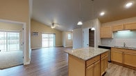 This single-floor space is defined by its striking vaulted ceilings, which lend an airy, expansive quality to the open-concept layout. The kitchen serves as a central hub, equipped with stainless steel appliances including a gas range, built-in microwave, dishwasher, and refrigerator. Light maple cabinetry lines the walls, complemented by a tile backsplash and granite countertops that extend across a central island, illuminated by pendant lighting overhead. The island provides additional seating and prep space, making it both functional and visually prominent.

The living area flows naturally from the kitchen, featuring wood-look flooring that carries throughout the main living spaces, a ceiling fan, and a sliding glass door that allows natural light to fill the room while offering outdoor access. Baseboard heating runs along the perimeter walls. A dedicated pantry provides ample storage adjacent to the kitchen.

The bathroom continues the cohesive design with matching maple cabinetry, granite-topped vanity, and wood-look vinyl flooring. Neutral warm-toned walls throughout create a consistent, welcoming atmosphere across the entire space.