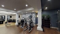 This single-level fitness center is thoughtfully designed to offer a complete workout environment within a stylish, modern setting. The space is immediately distinguished by a striking floor-to-ceiling typographic mural, featuring Los Angeles neighborhood names in bold black-and-white lettering, serving as a dynamic focal point throughout the room. White structural columns with classic detailing are positioned throughout, adding architectural character while naturally segmenting the space into distinct workout zones.

The cardio area is well-appointed with multiple elliptical trainers and treadmills, each equipped with digital displays. A dedicated strength training section houses a cable machine system, a rack of dumbbells arranged along mirrored walls, and additional resistance equipment. The flooring transitions between a warm herringbone wood-pattern and light-toned tile, delineating different functional areas effectively.

Recessed lighting throughout provides consistent, even illumination across all zones. Deep navy-blue accent walls contrast with the lighter portions of the room, contributing to a contemporary yet inviting atmosphere. Large floor-to-ceiling windows and a glass door along one wall open visually to a landscaped outdoor patio, introducing natural light and a connection to the exterior environment. Seating elements and greenery near the windows further enhance the overall ambiance.