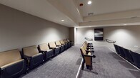 Located at 10977 Pacific Point Pl in San Diego, CA, this dedicated screening room delivers a polished, cinema-inspired experience on a single level. The space is anchored by a large wall-mounted flat-screen display set against a striking textured accent wall with a warm, natural finish, creating a compelling focal point for presentations or movie screenings. Rows of theater-style seats with tan upholstery and dark frames are arranged in a classic auditorium layout, each equipped with cup holders for added comfort. The seating is organized across two side-by-side sections, providing clear sightlines to the screen from every position. Patterned, commercial-grade carpeting in muted gray tones covers the floor throughout, contributing to the room's acoustic-friendly environment. Recessed lighting in the tray ceiling provides ambient illumination, while wall sconces and framed black-and-white landscape artwork lend a refined aesthetic to the neutral-toned walls. A vintage-style red popcorn machine stationed near the rear adds a charming, authentic movie-house touch. Tall decorative plants placed near the screen add a natural element to the room. The space is well-suited for private film screenings, community gatherings, or professional presentations.