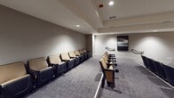 Located at 10977 Pacific Point Pl in San Diego, CA, this dedicated screening room delivers a polished, cinema-inspired experience on a single level. The space is anchored by a large wall-mounted flat-screen display set against a striking textured accent wall with a warm, natural finish, creating a compelling focal point for presentations or movie screenings. Rows of theater-style seats with tan upholstery and dark frames are arranged in a classic auditorium layout, each equipped with cup holders for added comfort. The seating is organized across two side-by-side sections, providing clear sightlines to the screen from every position. Patterned, commercial-grade carpeting in muted gray tones covers the floor throughout, contributing to the room's acoustic-friendly environment. Recessed lighting in the tray ceiling provides ambient illumination, while wall sconces and framed black-and-white landscape artwork lend a refined aesthetic to the neutral-toned walls. A vintage-style red popcorn machine stationed near the rear adds a charming, authentic movie-house touch. Tall decorative plants placed near the screen add a natural element to the room. The space is well-suited for private film screenings, community gatherings, or professional presentations.